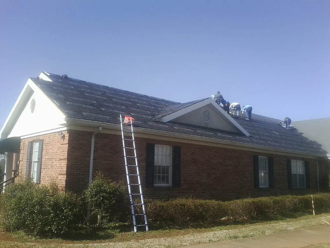 Crew tearing shingles off a brick ranch roof with a ladder set — Emergency Roof Repair work by our crew in Suwanee