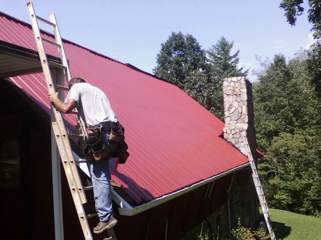 Roofer on a ladder beside a red metal roof and stone chimney — Soffit & Fascia Repair by Roofing Brothers in Suwanee